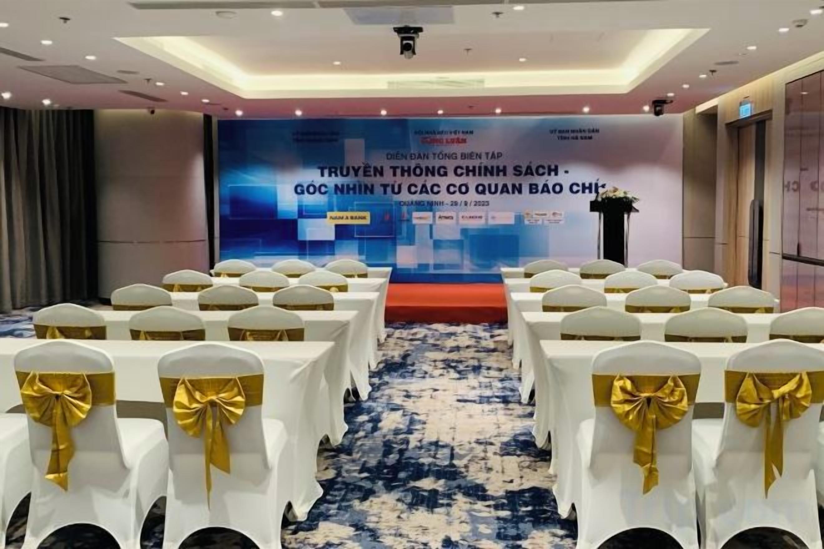 A conference room with rows of white chairs with gold sashes, a stage with a blue backdrop, and a carpeted floor.
