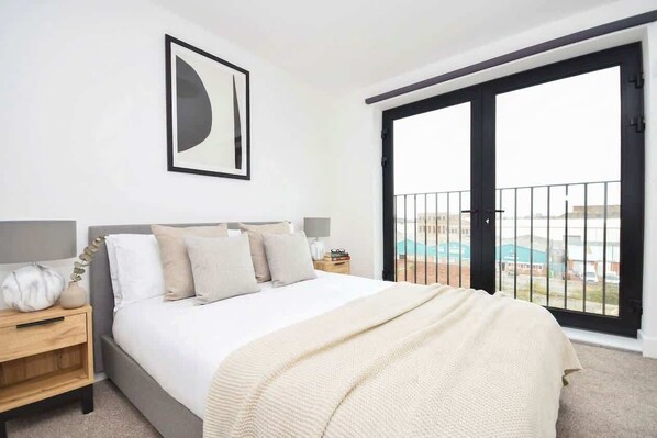 A bedroom with a bed, a nightstand, a lamp, a framed picture, and a balcony with a view.