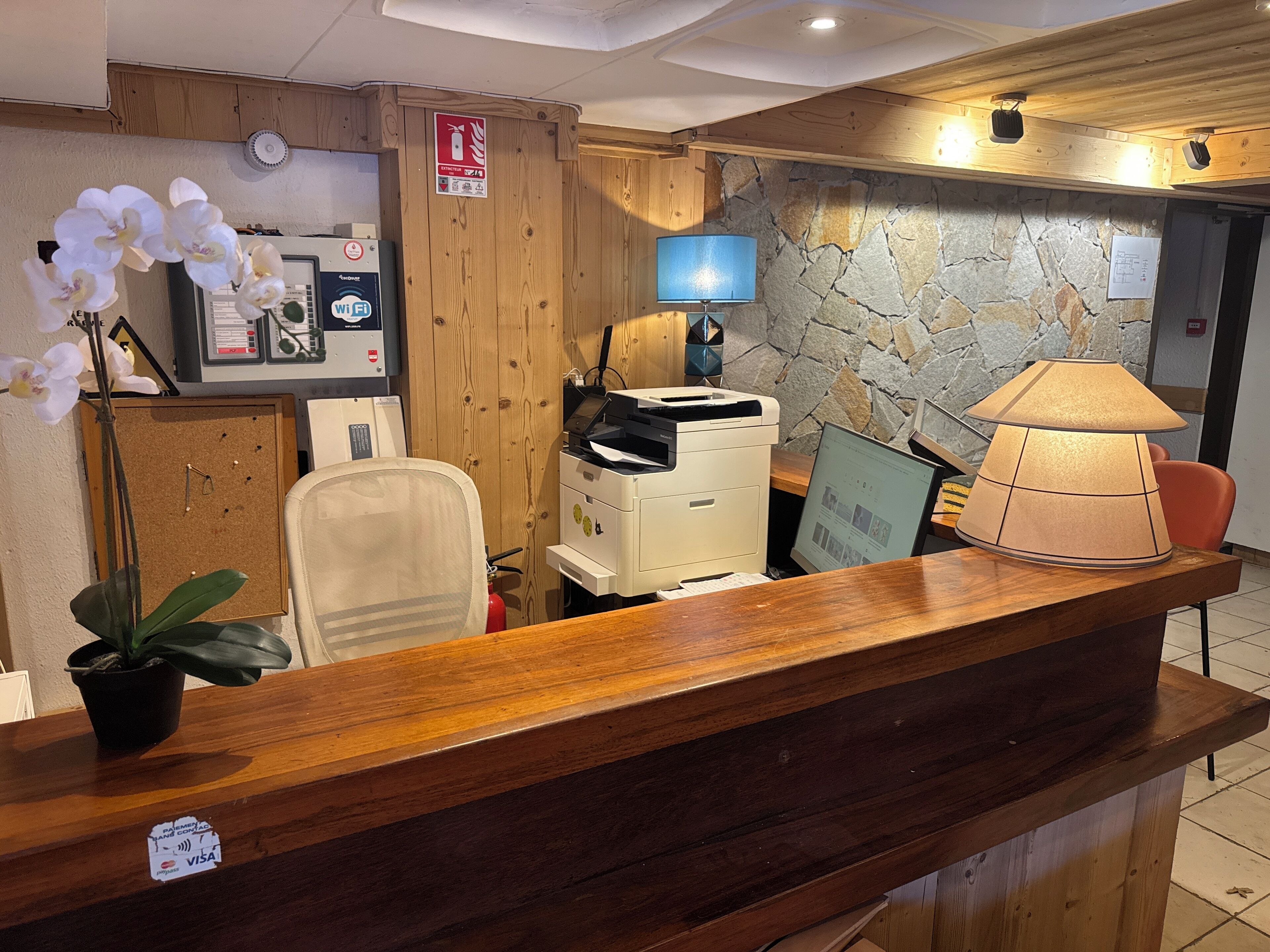 A reception desk with a wooden finish, a computer, a printer, and a lamp. There is a fire extinguisher and a sign on the wall.