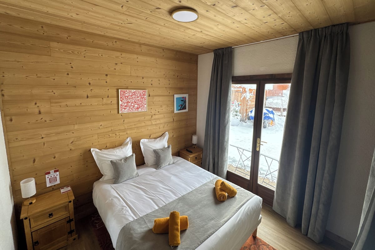 A wooden-paneled room with a bed, two pillows, a nightstand, a lamp, and a window with curtains.
