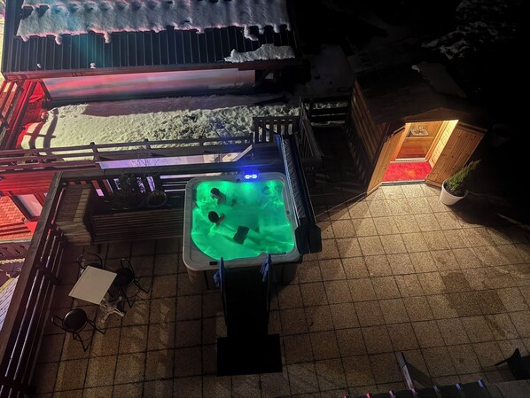 Un bain à remous où se trouvent deux personnes, une terrasse en bois et, au fond, un bâtiment avec un toit rouge.