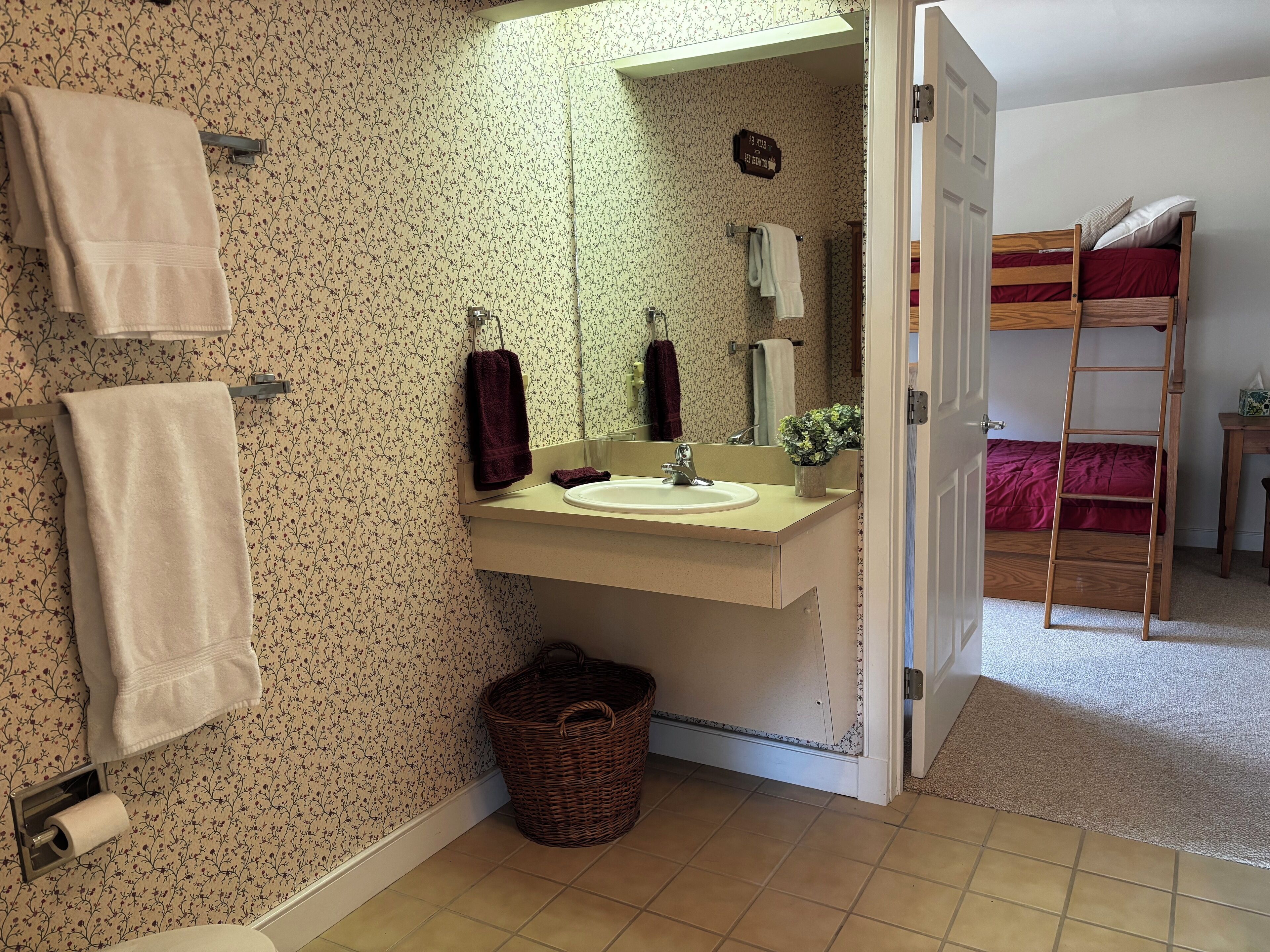 Ein Badezimmer mit blumigem Wandpapier, einem Waschbecken, einem Spiegel, einer Holzleiter und einem Stockbett im angrenzenden Raum.