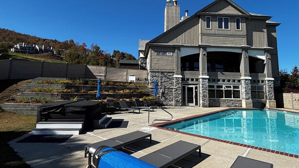 A pool area with lounge chairs and a covered grill, a stone facade building with a chimney, and a view of a hillside with houses.