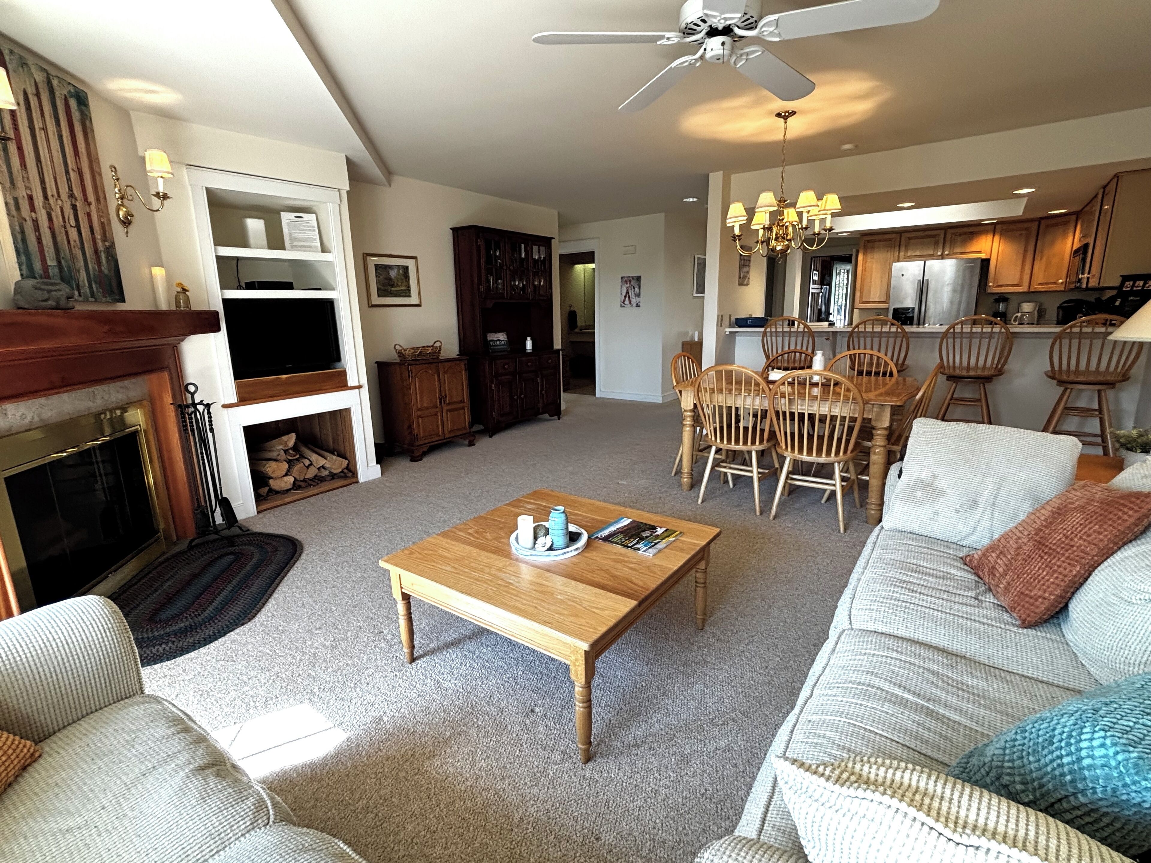 A spacious living room with a fireplace, a dining area with wooden chairs, and a kitchen in the background.
