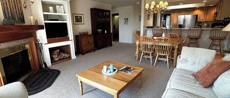 A spacious living room with a fireplace, a dining area with wooden chairs, and a kitchen in the background.