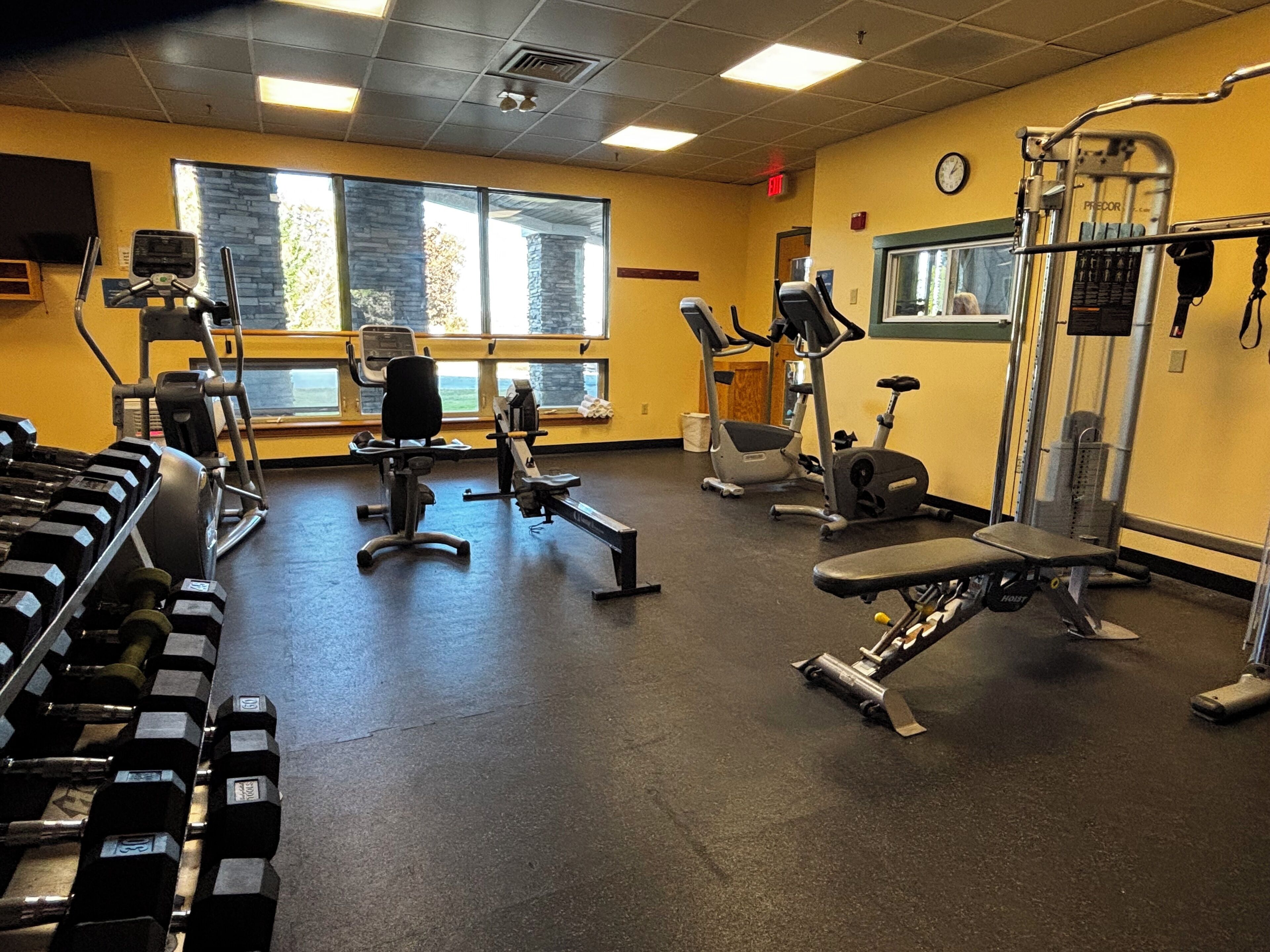 A gym with various exercise equipment, large windows, and a clock on the wall.