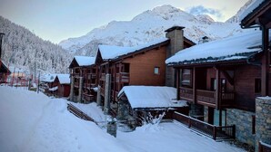 Un chalet de montagne enneigé, avec des cabanes en bois et des murs de pierre, se dressant sur un fond de sapins et de montagnes couverts de neige.