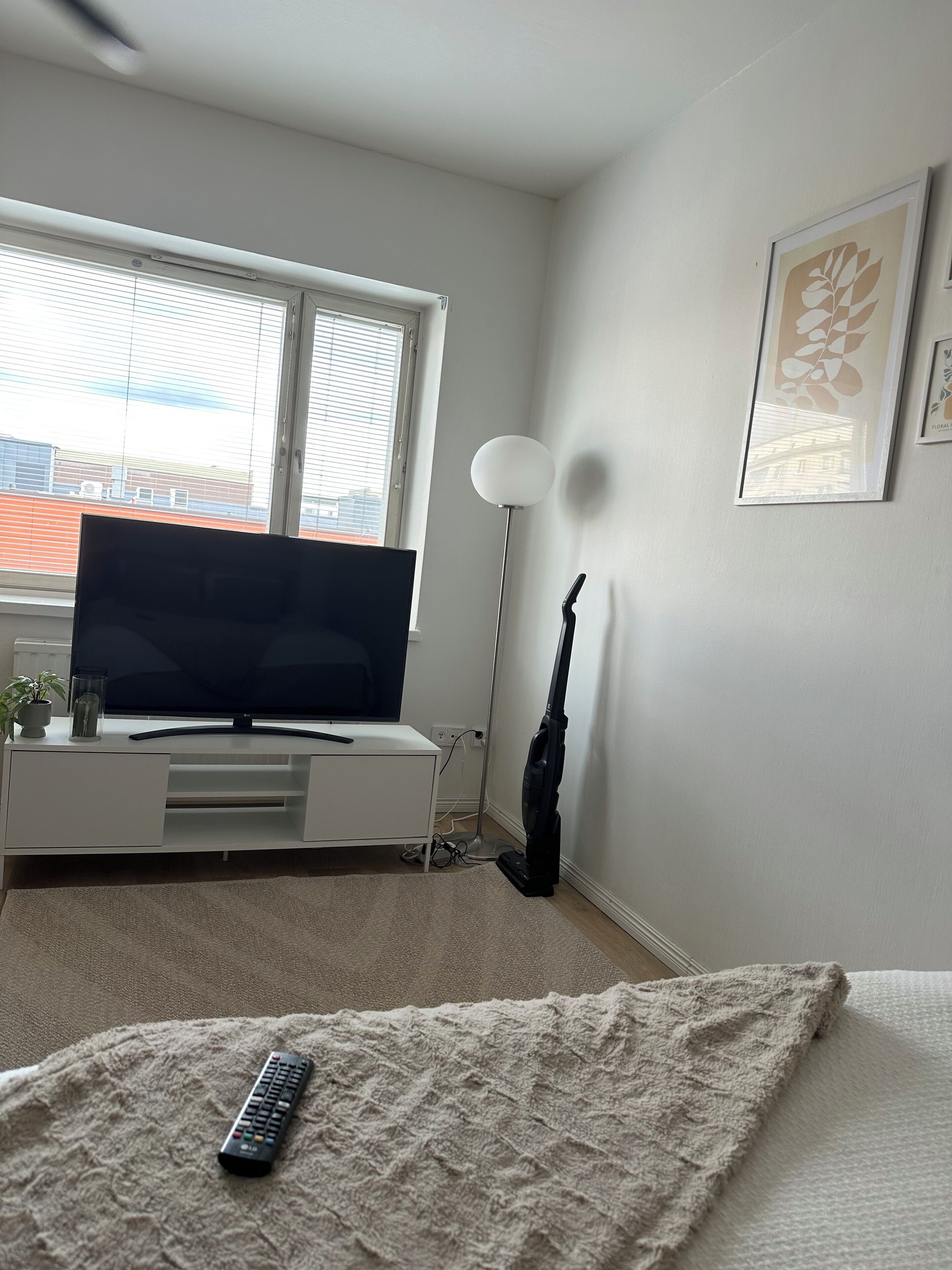Un dormitorio con una cama, un televisor sobre un mueble bajo, un aspirador y una ventana con persianas.