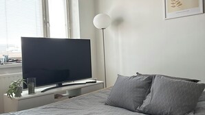 A bedroom with a bed, a flat-screen TV, a bedside table with a lamp, and a window with blinds. - Magnificent 1-bedroom apartment in Kouvola city centre (Kouvola)