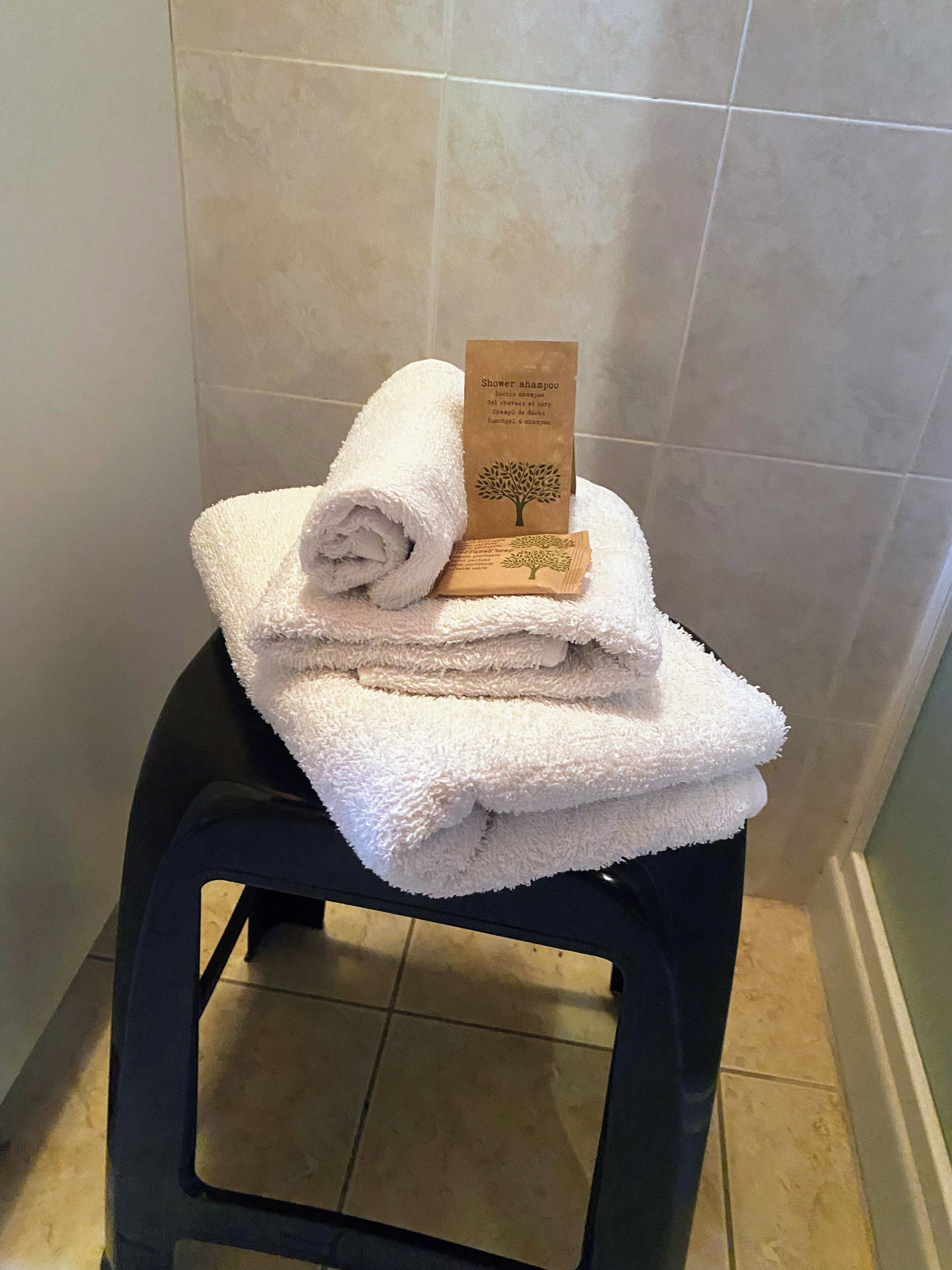 Une pile de serviettes blanches est posée sur un tabouret noir. Une étiquette marron indiquant « shampoing pour la douche » est fixée à côté. La salle de bain a des murs recouverts de carreaux.