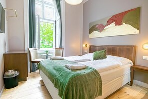 A hotel room with a bed, a chair, a small table, a painting, and a window with curtains.
