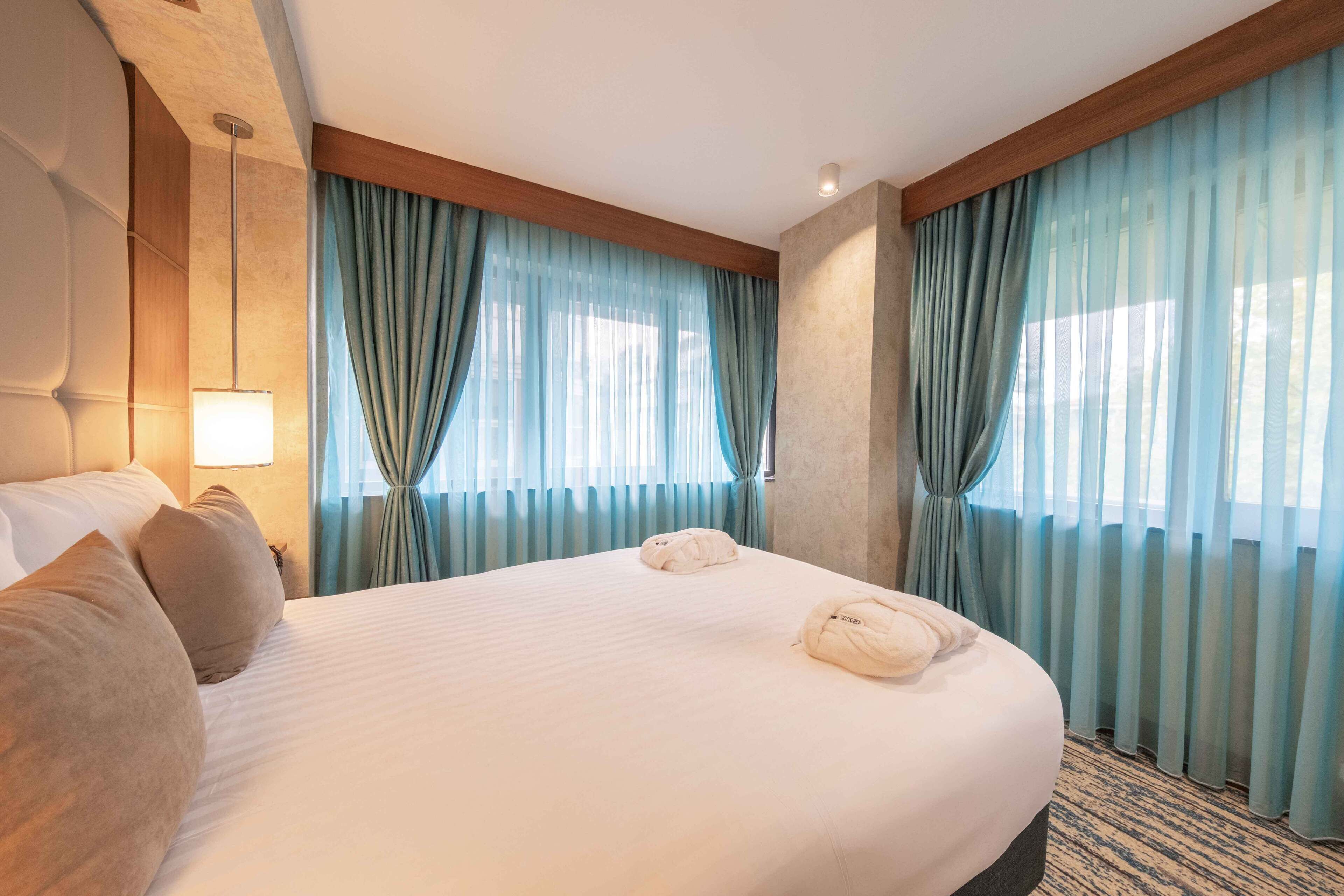 Una habitación de hotel con una cama grande, dos almohadas, una lámpara de noche y cortinas azul claro.
