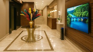 Un vestíbulo de hotel con suelo de mármol, una mesa redonda de cristal con un jarrón decorativo, una escalera, un mostrador de recepción y una gran pantalla de televisión mostrando un paisaje.