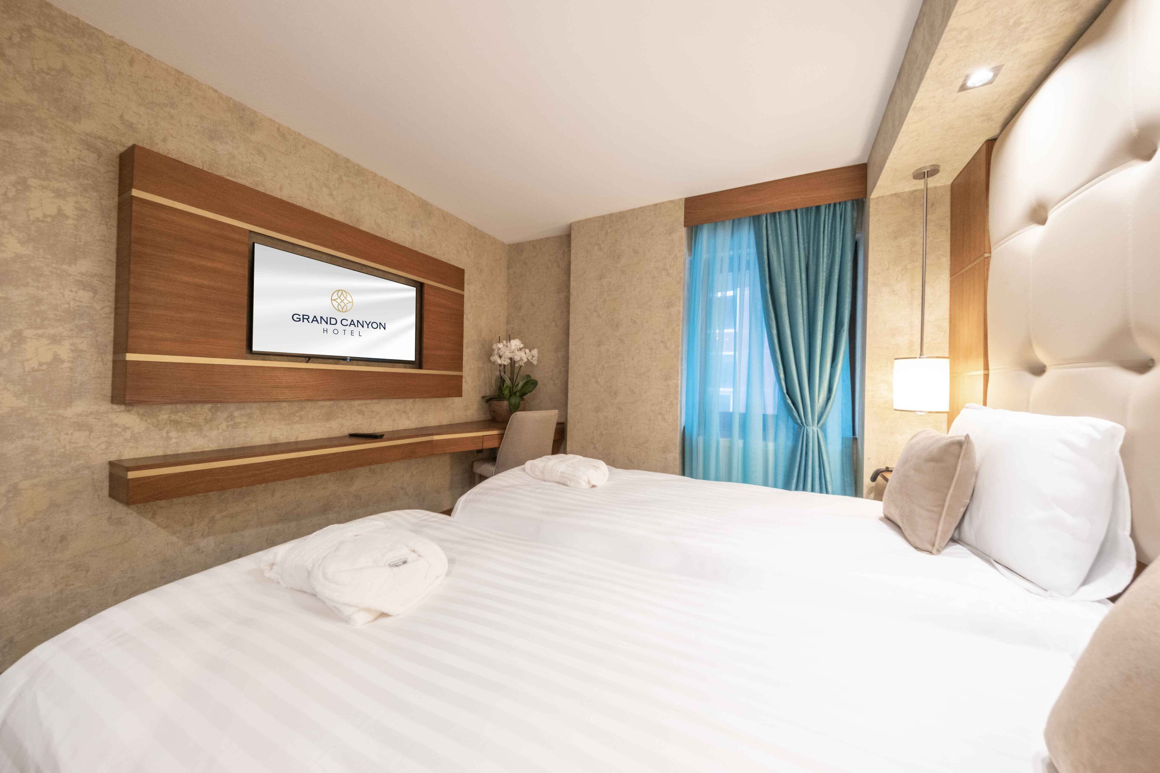 Una habitación de hotel con cama, televisor montado, mesita de noche y ventana con cortinas.