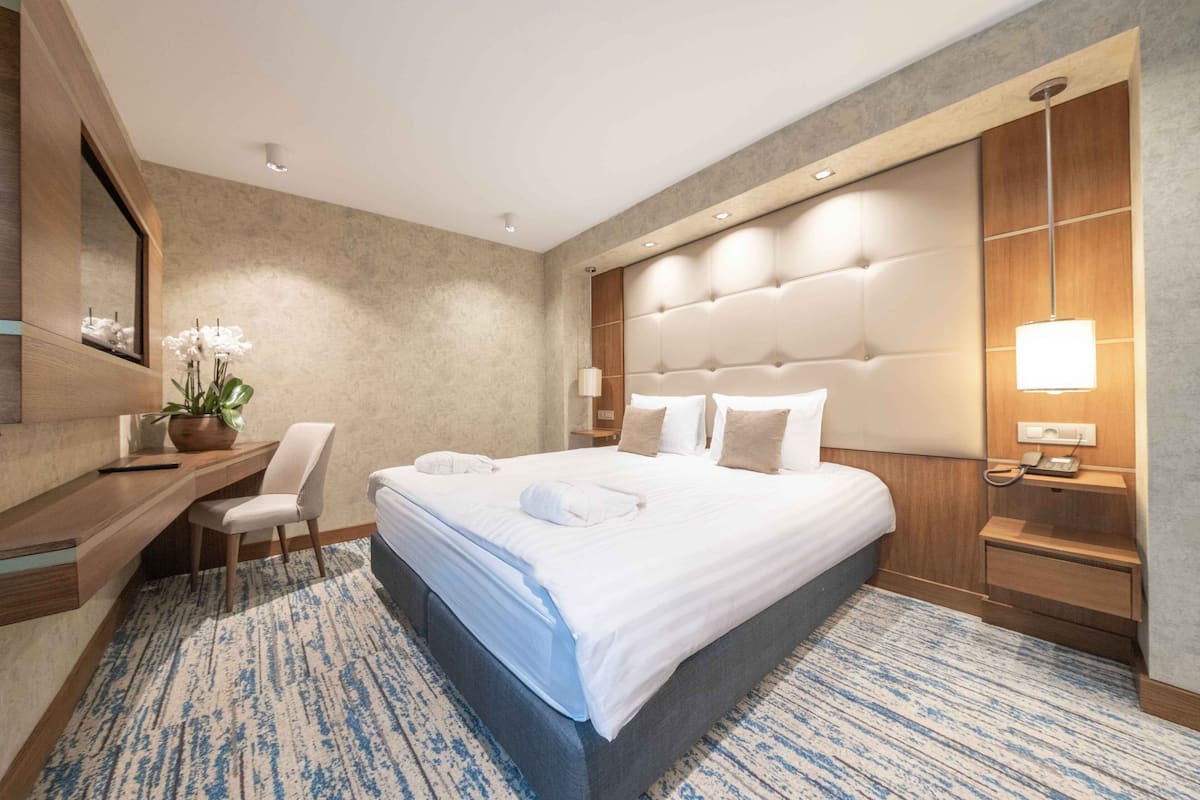 A modern hotel room with a large bed, a desk, a chair, and a bedside table with a lamp.