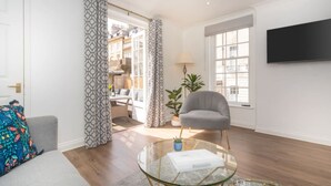 A modern living room with a glass coffee table, a grey armchair, and a TV mounted on the wall. - Crescent Mews Apartment (Bath)