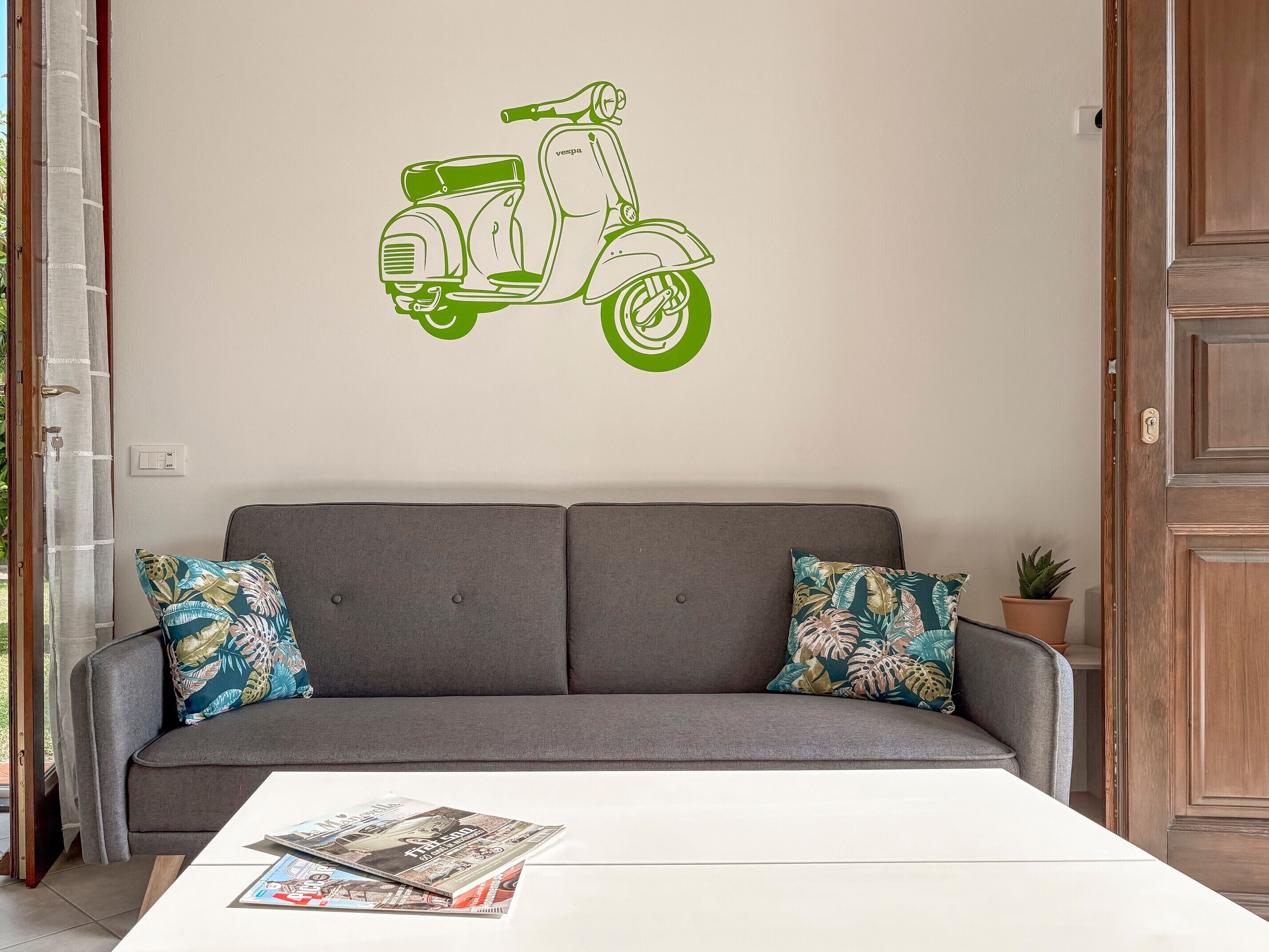 En grå sofa med mønstrede puder, et hvidt bord med magasiner, og en klistermærke af en scooter på væggen.