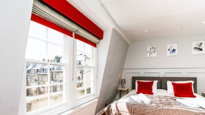 A bedroom with a large window, a bed with red pillows, and artwork on the wall. - Georgian Apartment near Royal Crescent (Bath and North East Somerset)