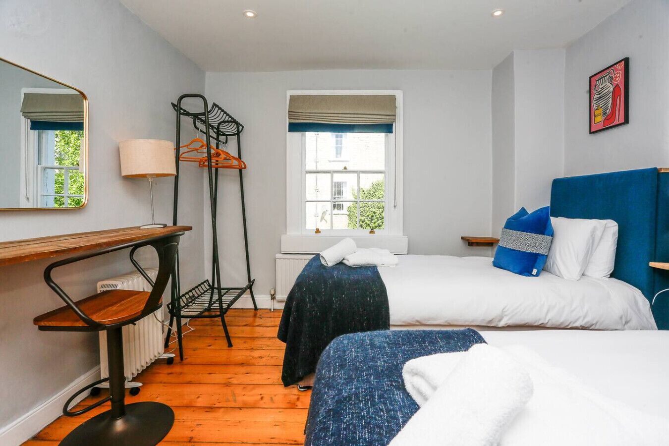 A bedroom with two beds, a radiator, a desk with a chair, a window with a view, and a wall-mounted shelf.