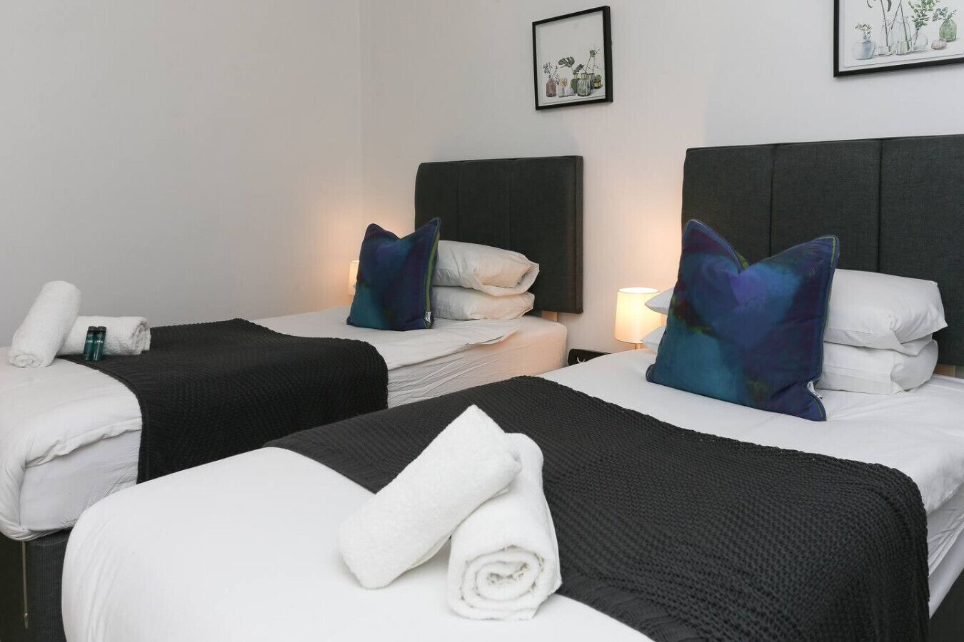 A hotel room with two beds, each with a black headboard, white linens, and a rolled towel. There is a framed picture on the wall above the beds.