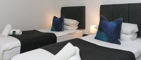 A hotel room with two beds, each with a black headboard, white linens, and a rolled towel. There is a framed picture on the wall above the beds.