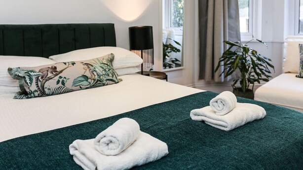 A neatly made bed with a green bedspread and two rolled towels, a pillow with a nature print, and a bedside table with a lamp. - Newbridge Road, Spacious One Bedroom (Bath)