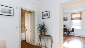 A hallway with wooden flooring, a door leading to a bathroom, a small table with a plant, and a dining area with chairs and a table. - Newbridge Road, Spacious One Bedroom (Bath)