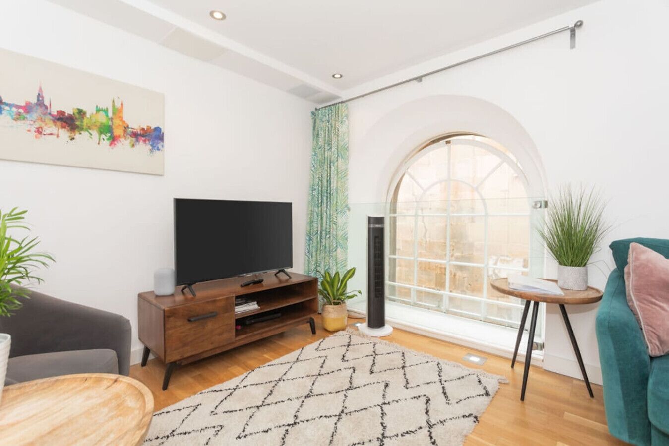 A modern living room with a flat-screen TV, wooden entertainment unit, a patterned rug, a teal armchair, and a large arched window with a view.
