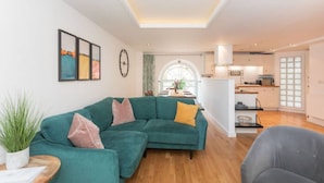 Una sala de estar moderna con un sofá color turquesa, un sillón gris y una mesa de centro de madera. Se ve una cocina al fondo, con armarios blancos y un ventanal grande.