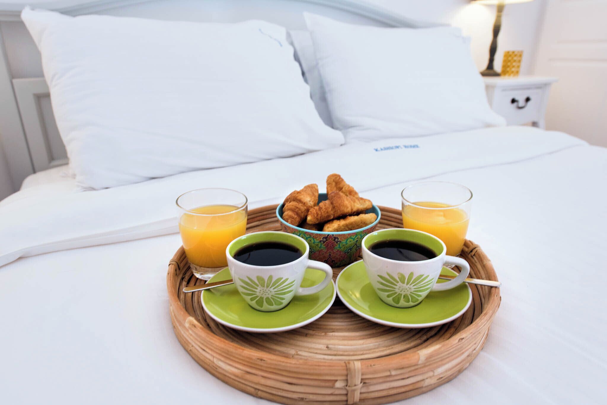 Een ontbijfwagen met croissants, koffie en sinaasappelsap op een bed met witte lakens en kussens.