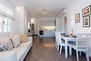 Una cocina moderna con armarios blancos, una mesa de comedor con sillas blancas y un sofá de color claro con cojines estampados.