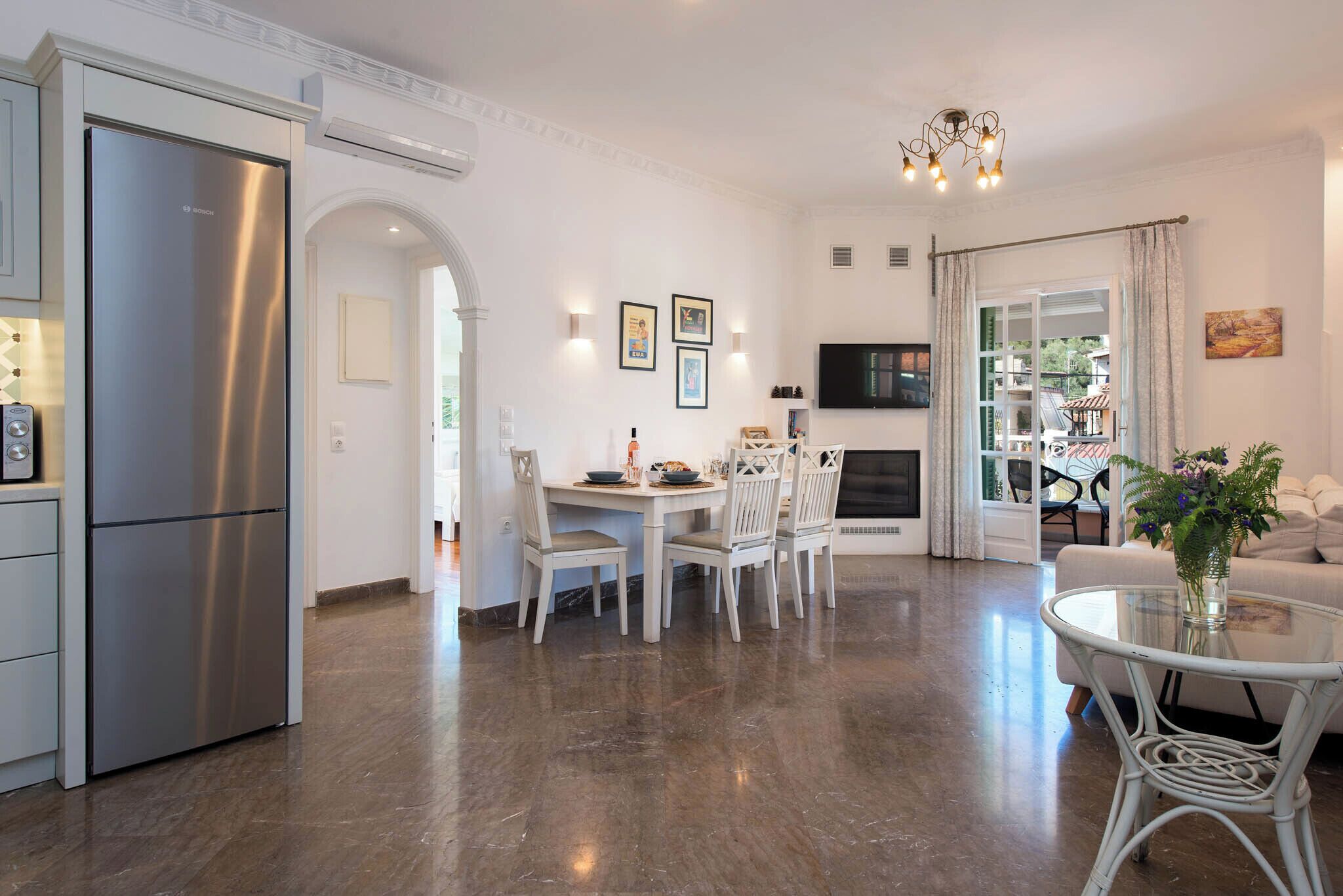 A modern kitchen with a stainless steel refrigerator, a dining area with white chairs and a table, and a living room with a sofa and a TV.