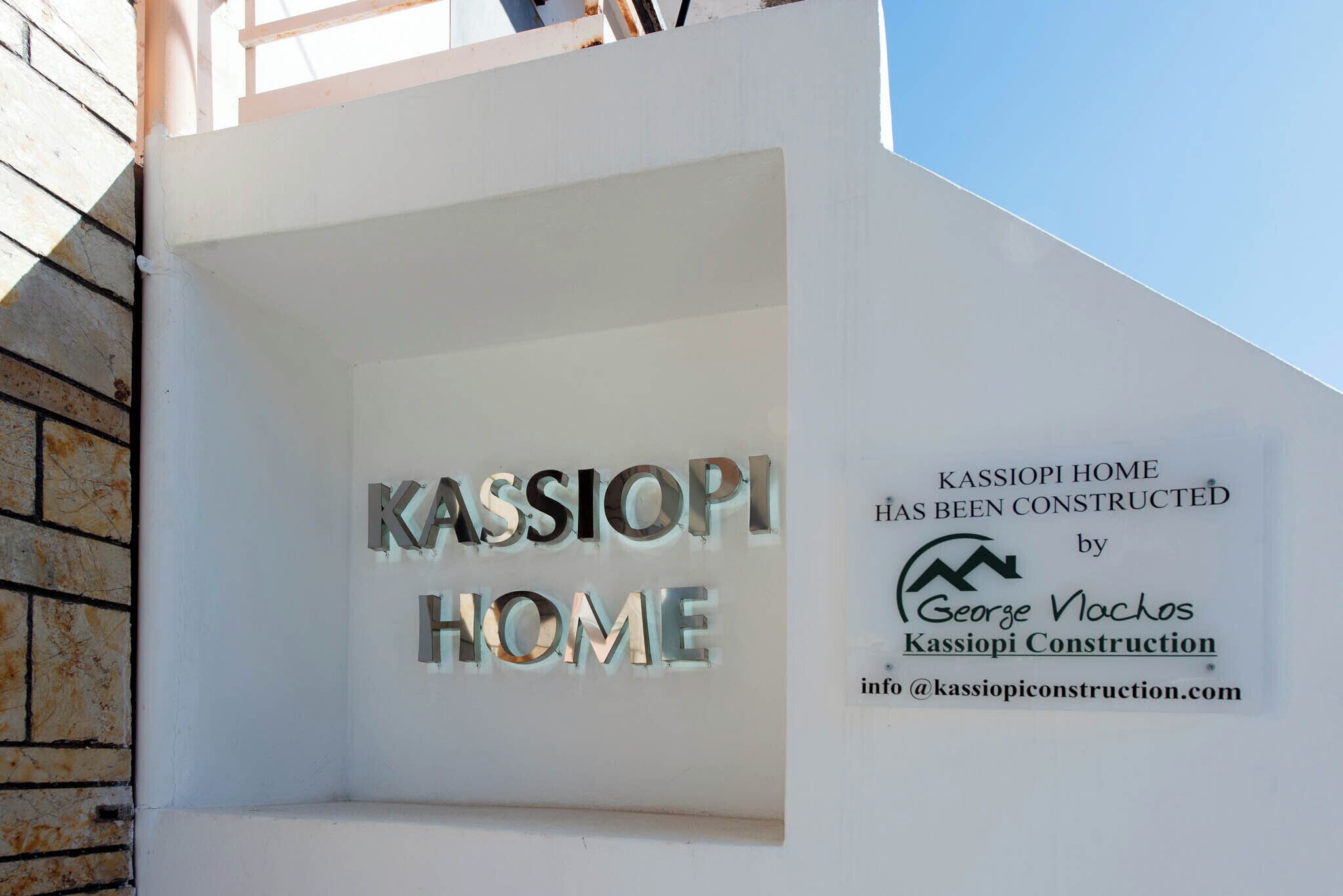 En vit vägg med texten ”KASSIOPI HOME” och en skylt med texten ”KASSIOPI HOME HAR BYGGTS av George Vochos, Kassipi Construction”.