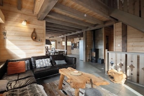 Een ruime woonkamer met een houten plafond, een zwarte leren bank, een houten salontafel en een open haard.