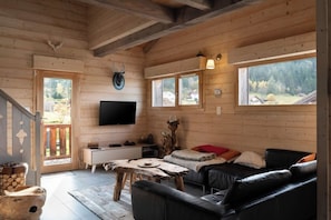 Interior de una cabaña de troncos con techo de madera, un televisor de pantalla plana montado en la pared, un sofá de cuero negro y una mesa de comedor rústica.