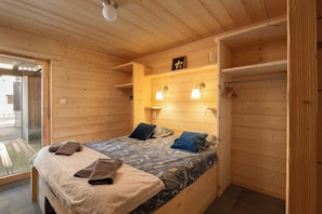 Habitación de cabaña de madera con una cama, un estante pequeño con luces y una puerta corrediza a un balcón.