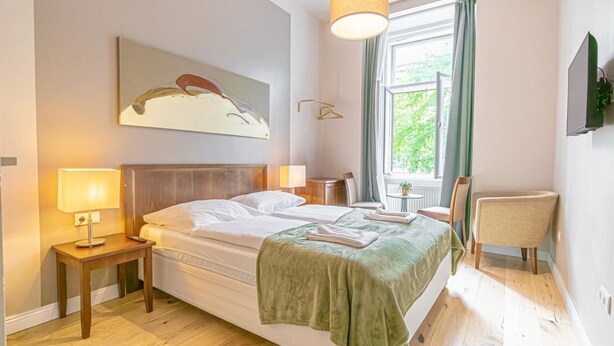 A hotel room with a bed, bedside lamps, a chair, a window with curtains, a painting on the wall, and a television mounted on the wall. - Chic Room with Private Bath | Mariahilferstraße (Vienna)
