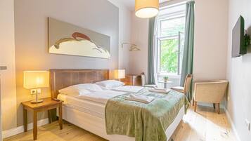 A hotel room with a bed, bedside lamps, a chair, a window with curtains, a painting on the wall, and a television mounted on the wall.