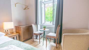 A hotel room with a bed, a small table with chairs, a radiator, and a window with curtains.