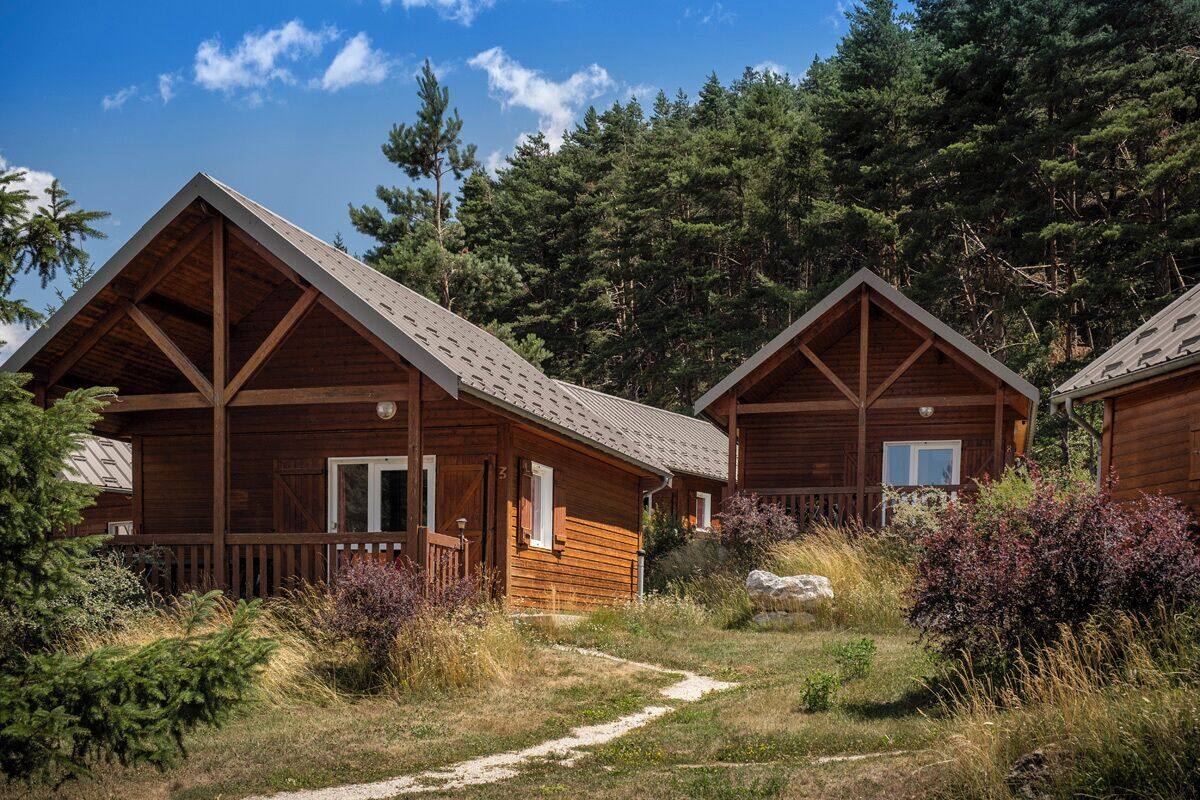 Two wooden cabins with pitched roofs, surrounded by a forested area and a gravel path.