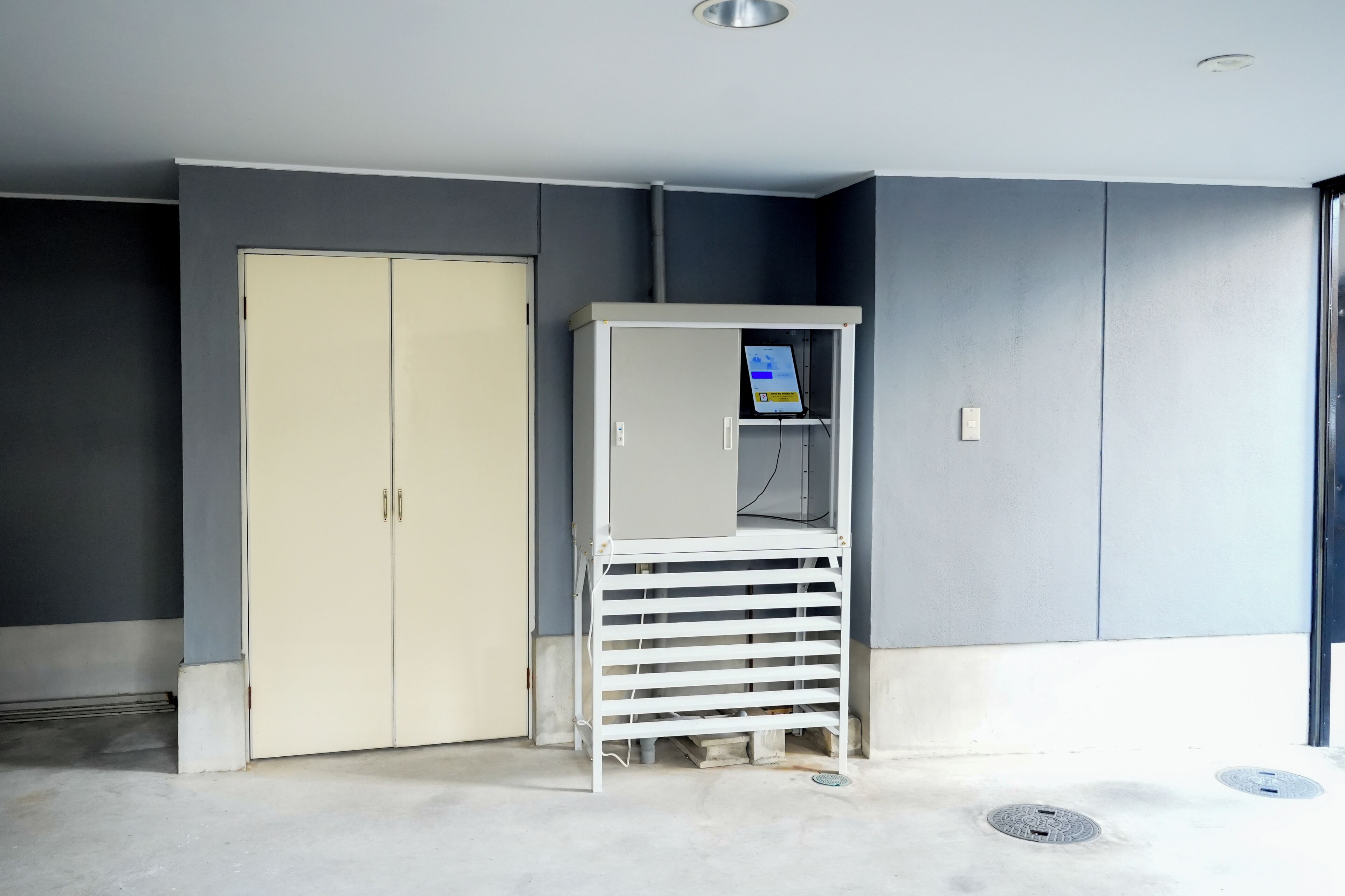 A room with a beige wardrobe, a white shelving unit with a control panel, and a grey wall.