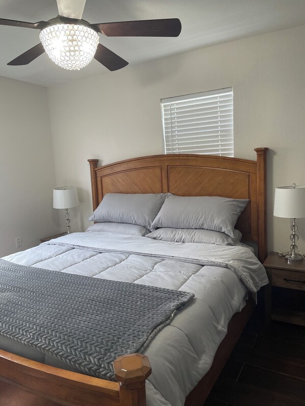 A bedroom with a wooden bed frame, two bedside tables with lamps, a ceiling fan, and a window with blinds.