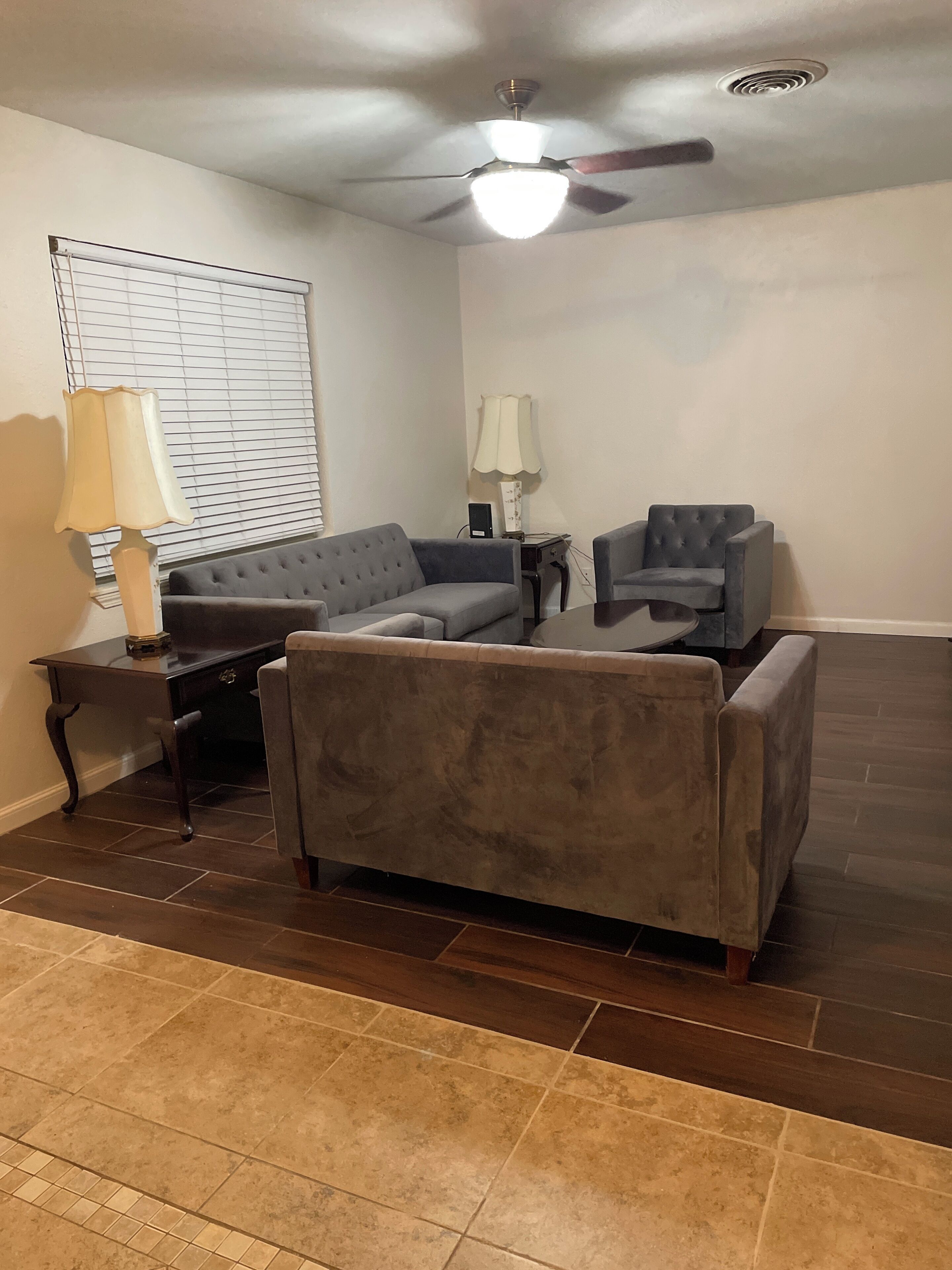 A living room with a grey sofa, a matching armchair, a small table, a lamp, and a ceiling fan.