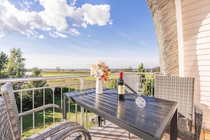 Un balcon avec une table, des chaises en osier, une bouteille de vin et un vase de fleurs.