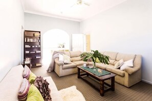 A living room with a sofa, coffee table, bookshelf, and a large window. - Aussie Chillout - Coal d' Vine Cottage - Cessnock NSW (Cessnock)