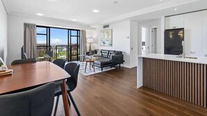 Ein modernes Wohnzimmer mit einer Couch, einem Esstisch und Stühlen. Es gibt eine Küchenzeile mit einem Kühlschrank und einem Fenster mit Blick auf eine Stadtlandschaft.