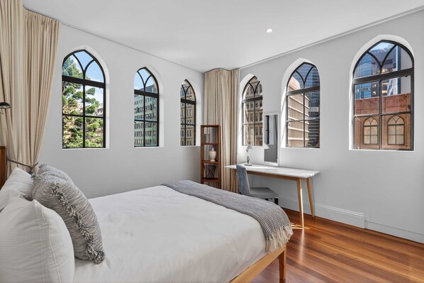 Ein Schlafzimmer mit einem großen Bett, einem Schreibtisch und hohen Bogenfenstern mit Blick auf Gebäude.
