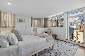 Ein Schlafzimmer mit Bett, Sessel, Fenster und Blick auf Gebäude.