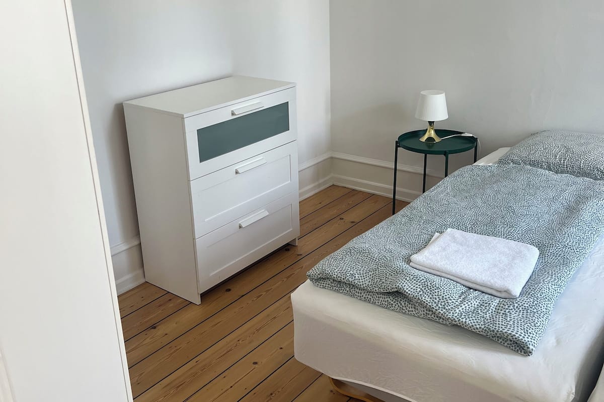 Un dormitorio con suelo de madera, una mesita de noche blanca con lámpara, una cama con manta estampada y un armario blanco.