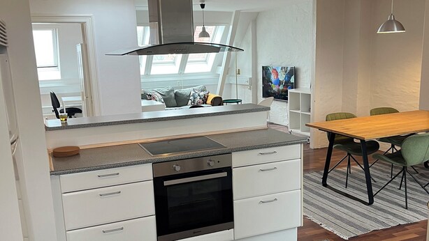 Eine moderne Küche mit weißen Schränken, einem eingebauten Backofen und einer Edelstahl-Dunstabzugshaube. Ein Essbereich mit einem Holztisch und grünen Stühlen ist ebenfalls vorhanden.